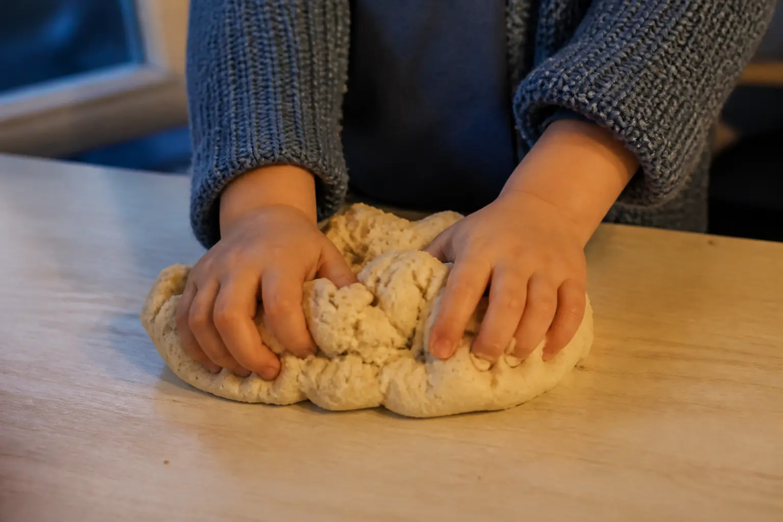 Mains pétrissant de la pate a modeler maison sur une table de cuisine, lumière chaude près d’une fenêtre