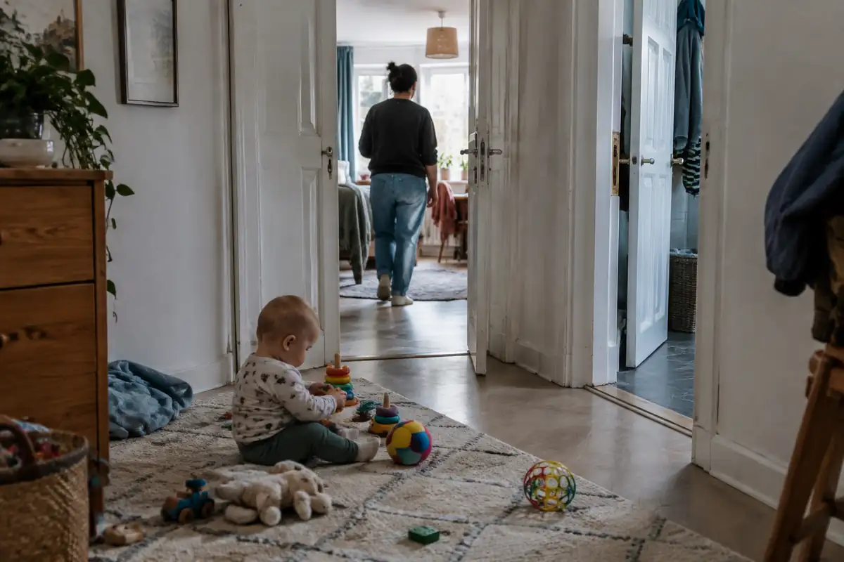Bébé qui joue sur un tapis pendant qu'un adulte, vu de dos, passe dans la pièce voisine