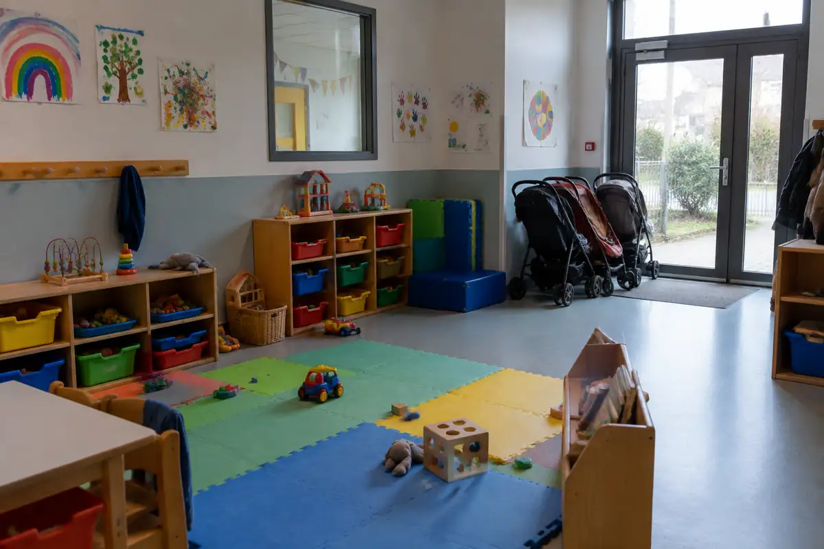 Salle d'éveil d'une crèche avec tapis de sol, étagères de jouets et poussettes garées à l'entrée